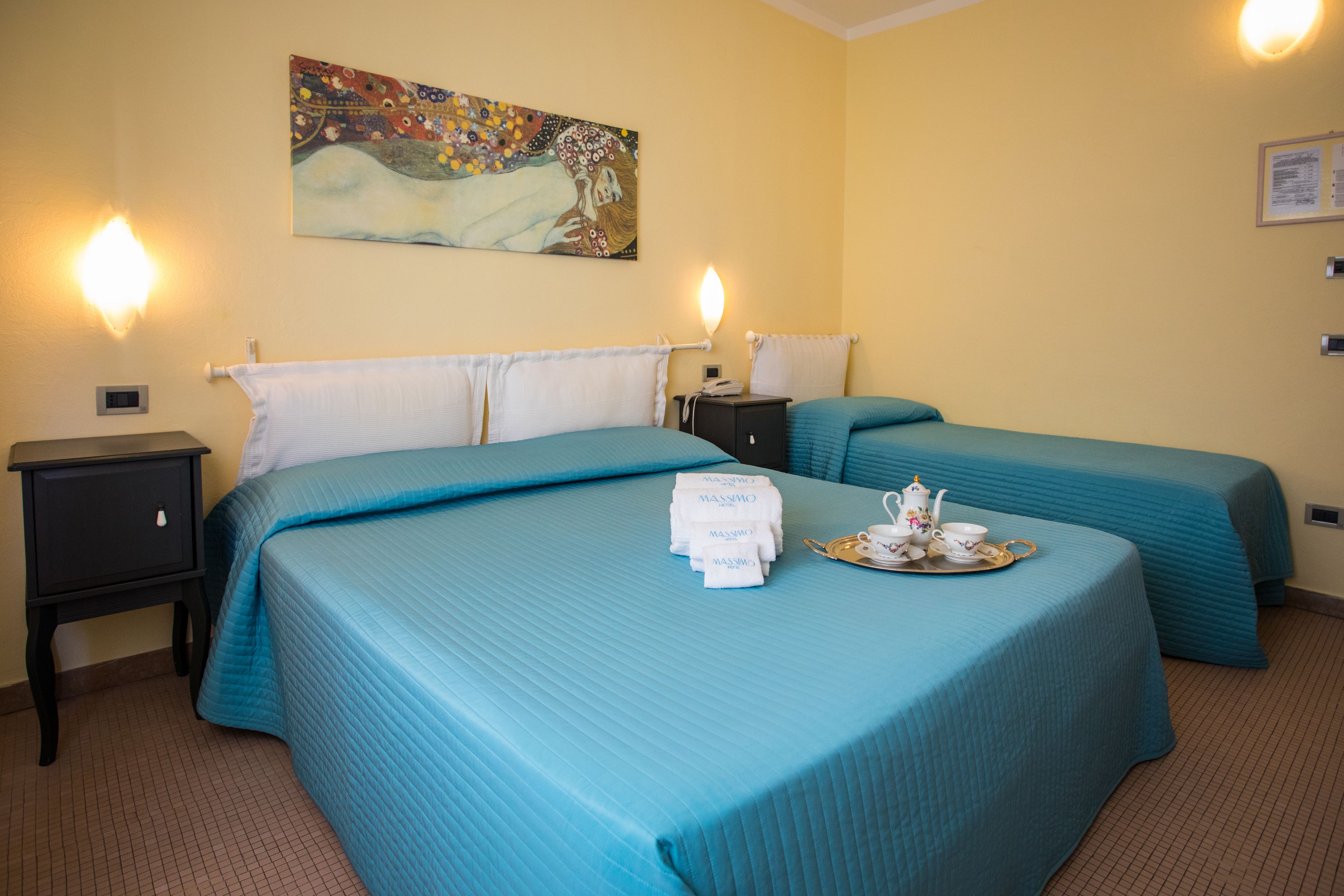 A hotel room with two beds, each with a blue bedspread and white pillows. There is a painting on the wall above the beds, a nightstand with a phone and a tray with a teapot and cups on the bed.