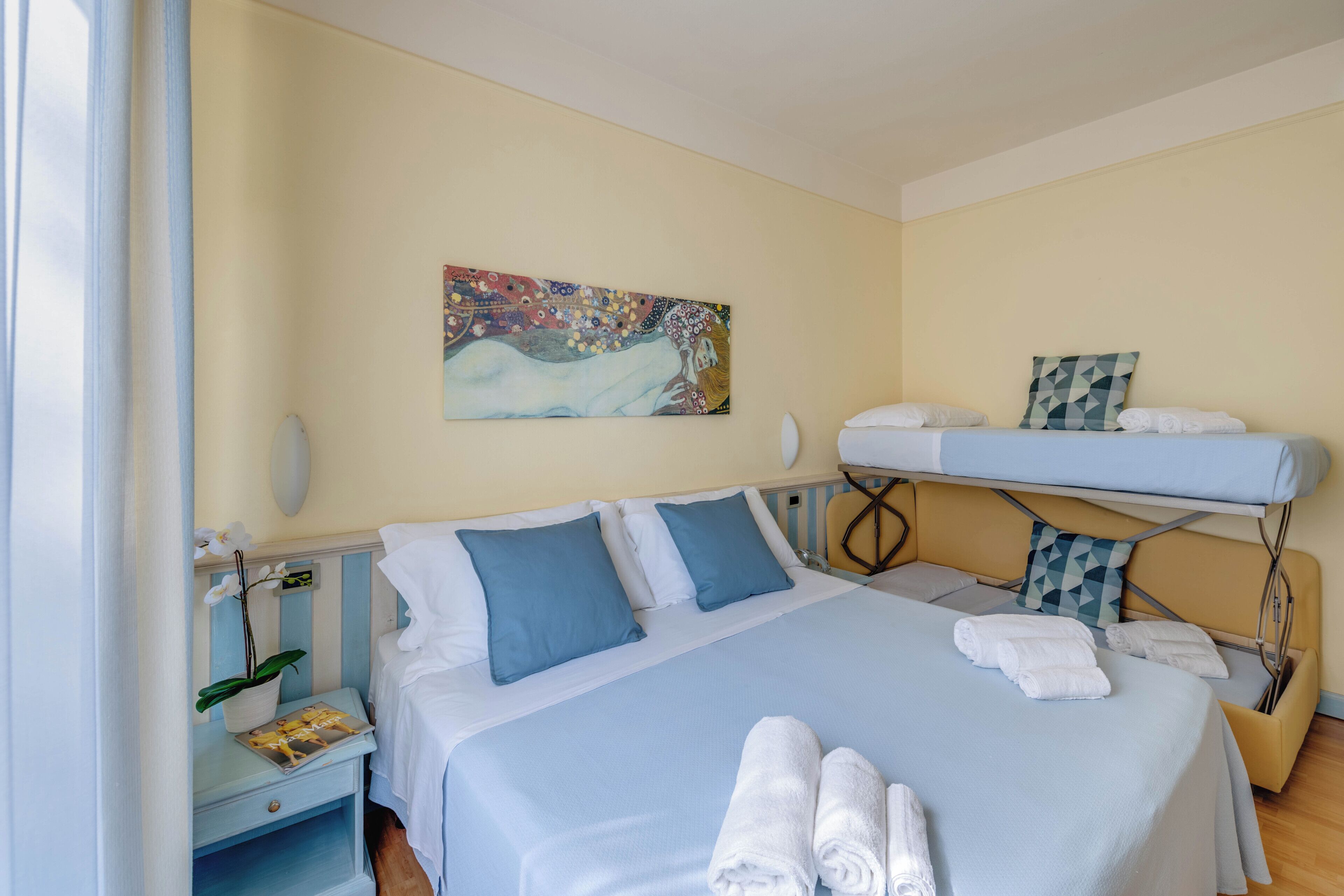 A hotel room with a bed, a nightstand, a painting, and a bunk bed.