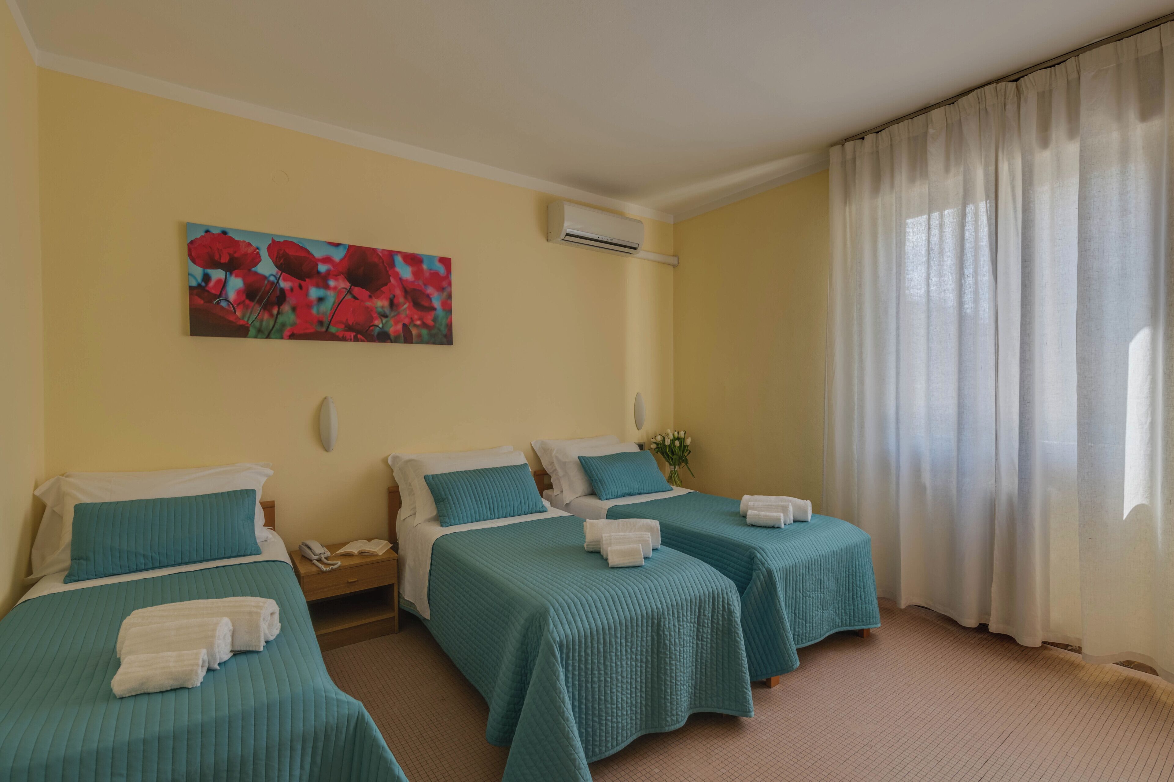 A hotel room with two beds, each with teal bedding and white pillows. There is a painting of red flowers on the wall, an air conditioning unit, and a window with sheer curtains.