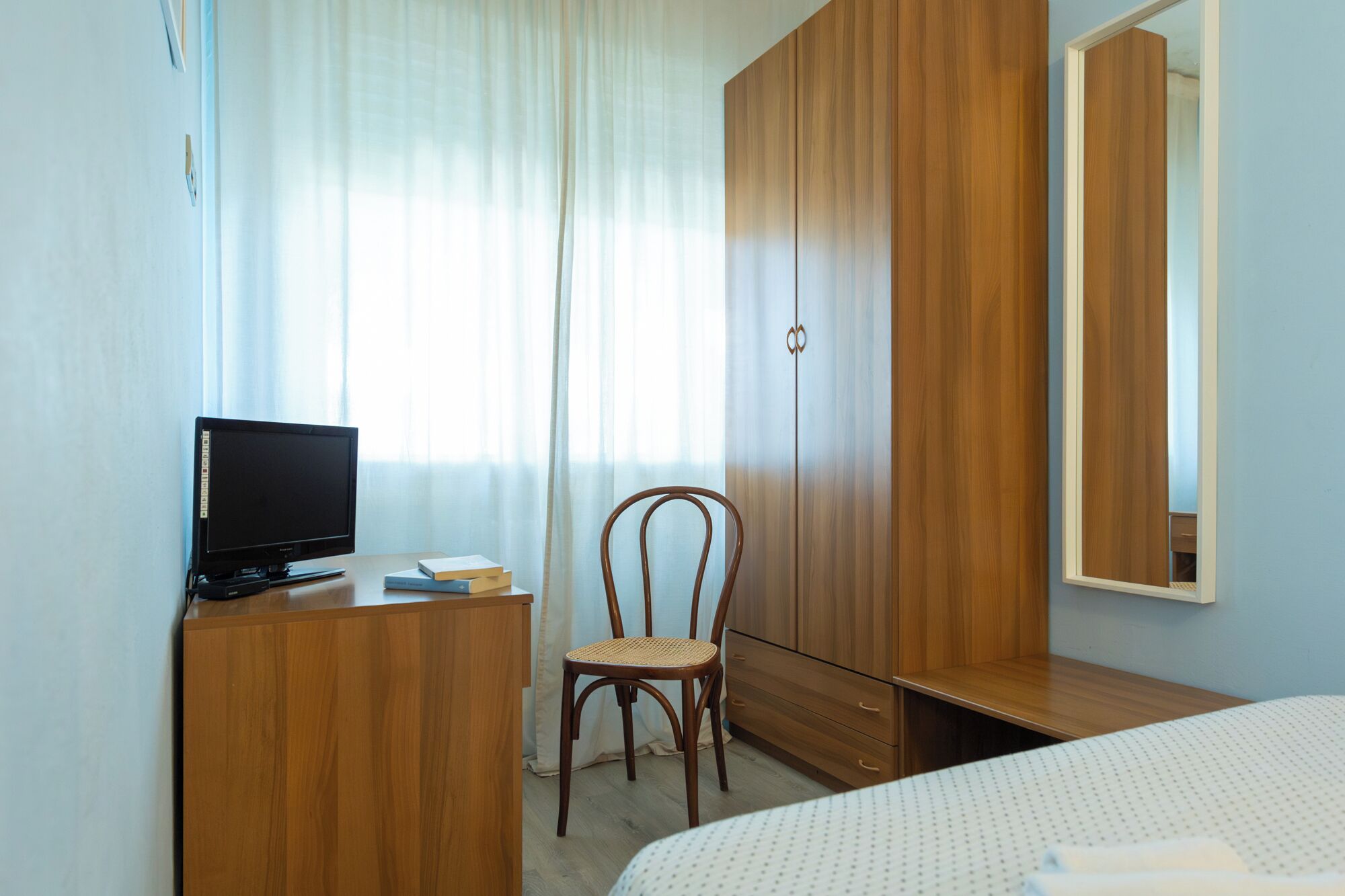A bedroom with a wooden wardrobe, a desk with a television, a chair, and a bed.