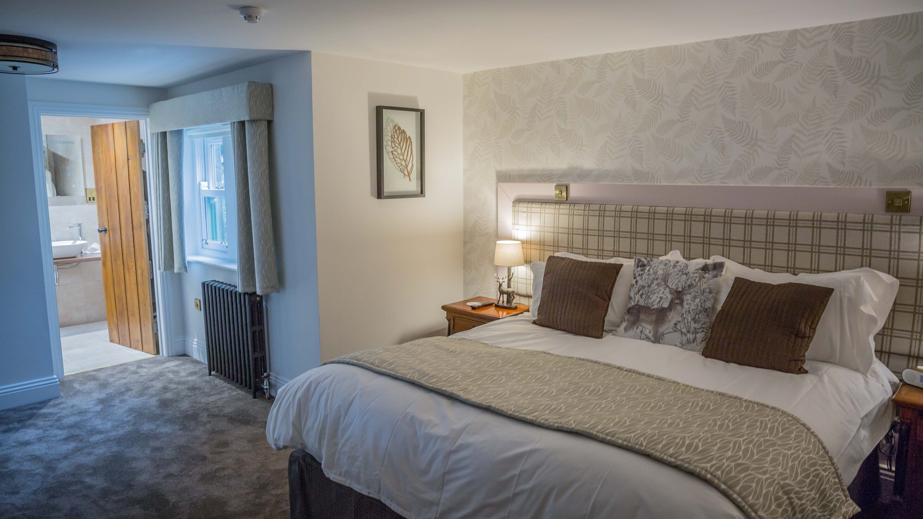 Una habitación de hotel con una cama grande, una mesita de noche con lámpara, un cuadro en la pared y una puerta que da a un baño.