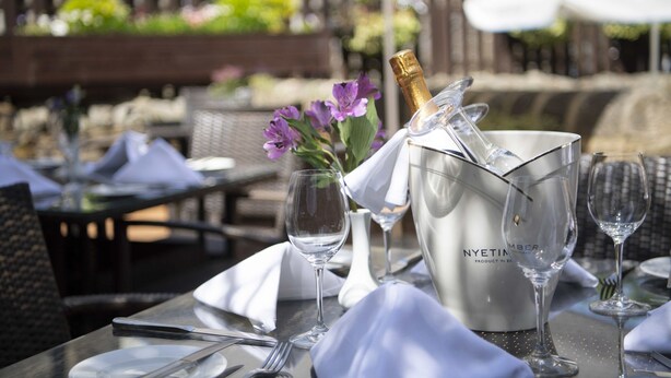 Un área de comedor al aire libre con una mesa preparada para dos personas, que incluye una botella de champán, copas de vino y un florero con flores moradas.