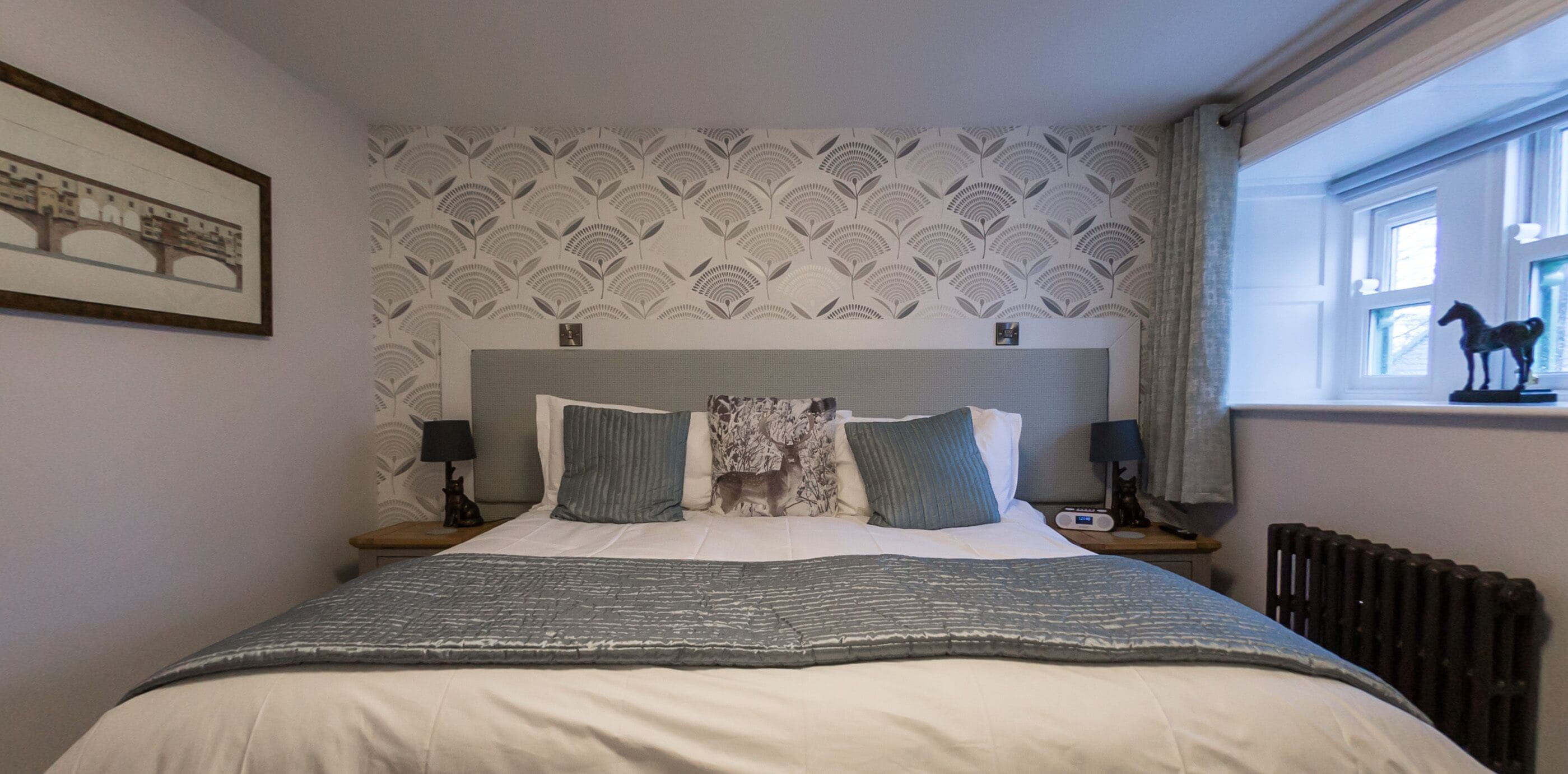 Una cama con arreglos cuidados, cabecera gris y blanca con diseño, dos mesitas de noche con lámparas, un radiador y una ventana con una estatua de un caballo.