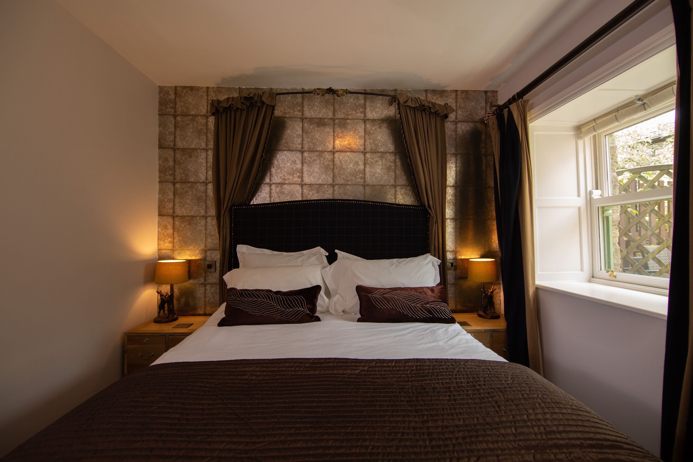 A neatly made bed with a headboard, two bedside tables with lamps, a window with curtains, and a stone-patterned wall.