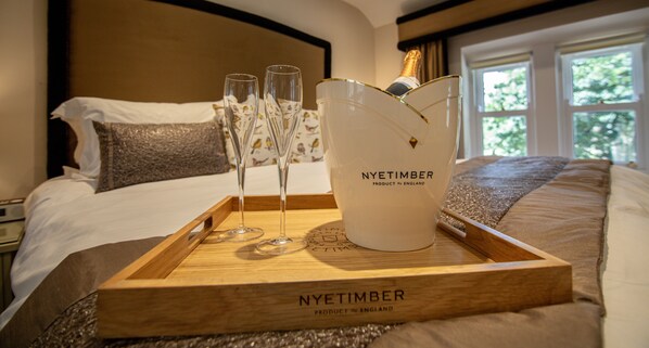 Una bandeja de madera con una botella de champán Nyetimber y copas sobre una cama, con vistas a árboles a través de la ventana.