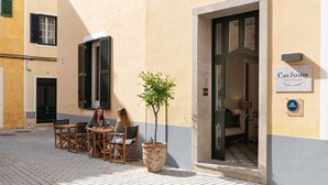 A café with outdoor seating, a potted tree, and a hotel entrance with a sign. - Hotel Boutique Can Sastre (Ciutadella de Menorca)