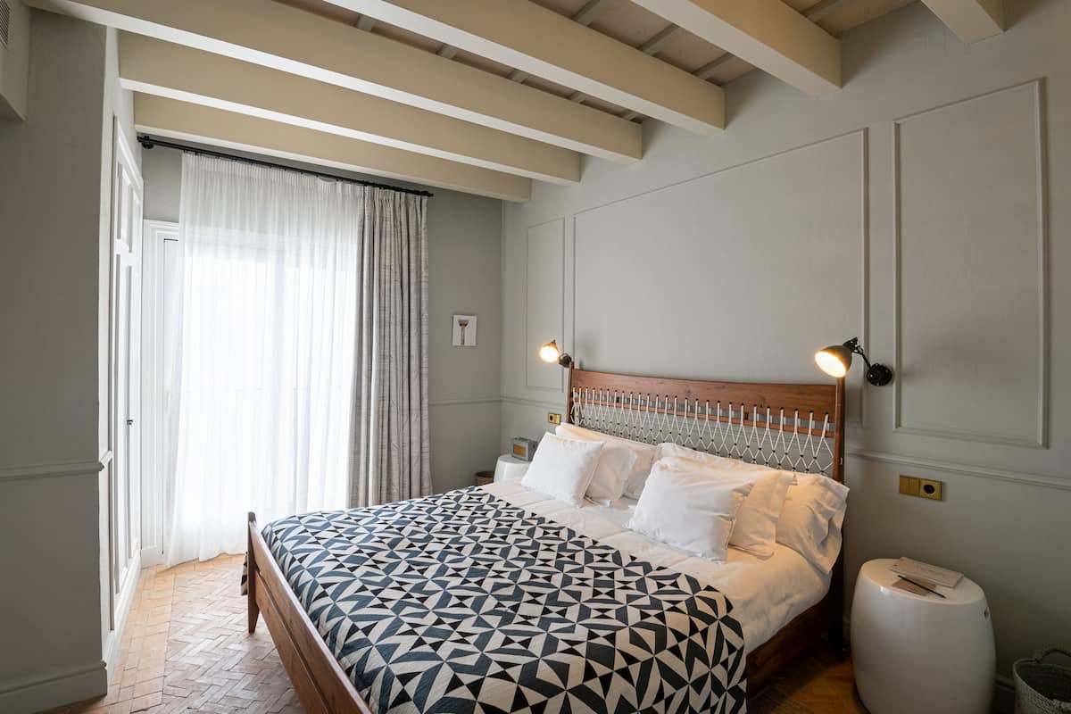 Une chambre avec un lit en bois, du linge de lit blanc, un tapis à motifs chevrons, une table de chevet avec une lampe et une fenêtre avec des rideaux légers.