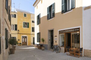 Una calle estrecha con un café llamado "PASTELLA LA SINNOMBRE CONFEITTERIA" a la derecha, con mesas al aire libre y una bicicleta estacionada afuera.