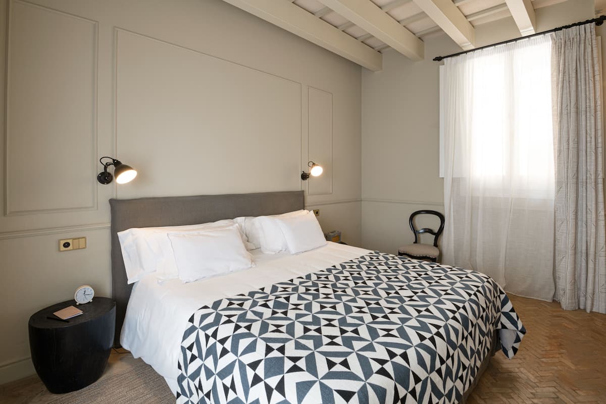 Une chambre à coucher comprenant un lit, une petite table ronde, une table de chevet, une chaise et une fenêtre avec des rideaux.