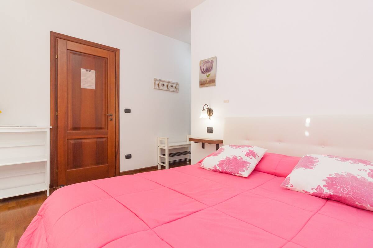 A bedroom with a wooden door, a white shelf, a bedside table with a lamp, and a bed with pink bedding.
