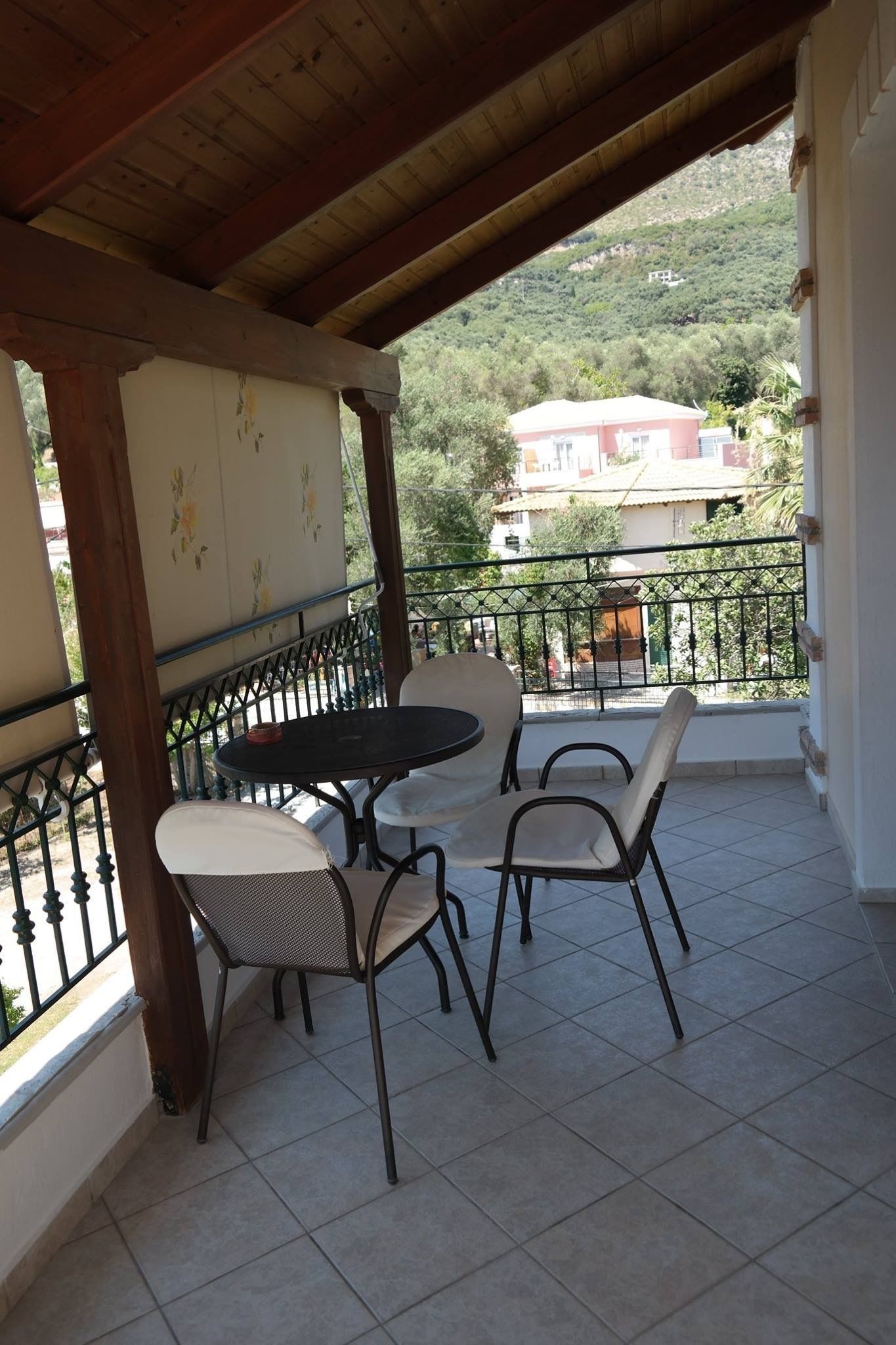 A covered balcony with a table and chairs, a view of a hillside with buildings, and a metal railing.