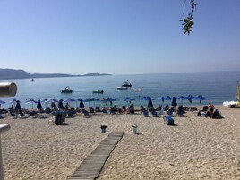 Una playa con tumbonas y sombrillas, un paseo marítimo de madera y barcos en el agua.