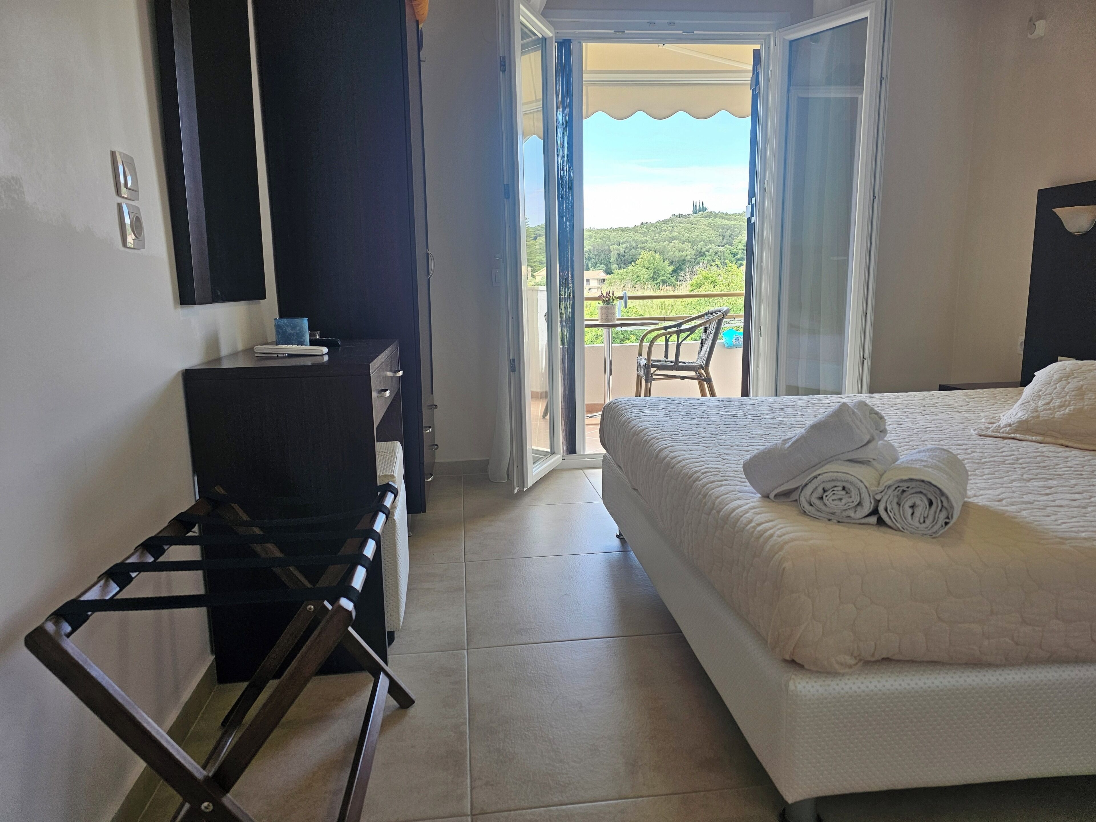 A hotel room with a bed, a desk with a chair, a TV, and a balcony with a view of the countryside.