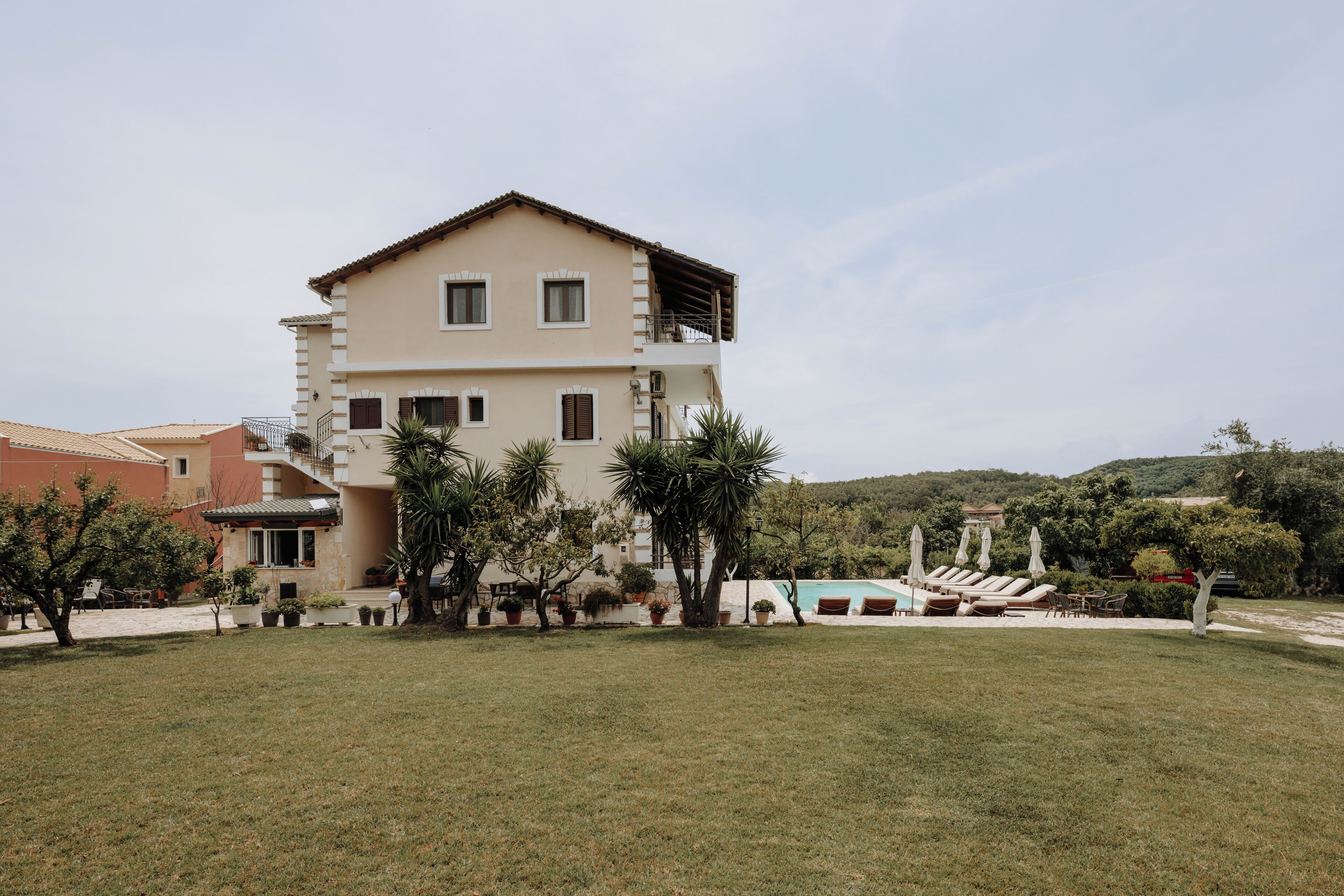 A two-story building with a flat roof, a balcony on the upper floor, and a swimming pool with sun loungers in the background.