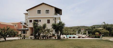 A two-story building with a flat roof, a balcony on the upper floor, and a swimming pool with sun loungers in the background.