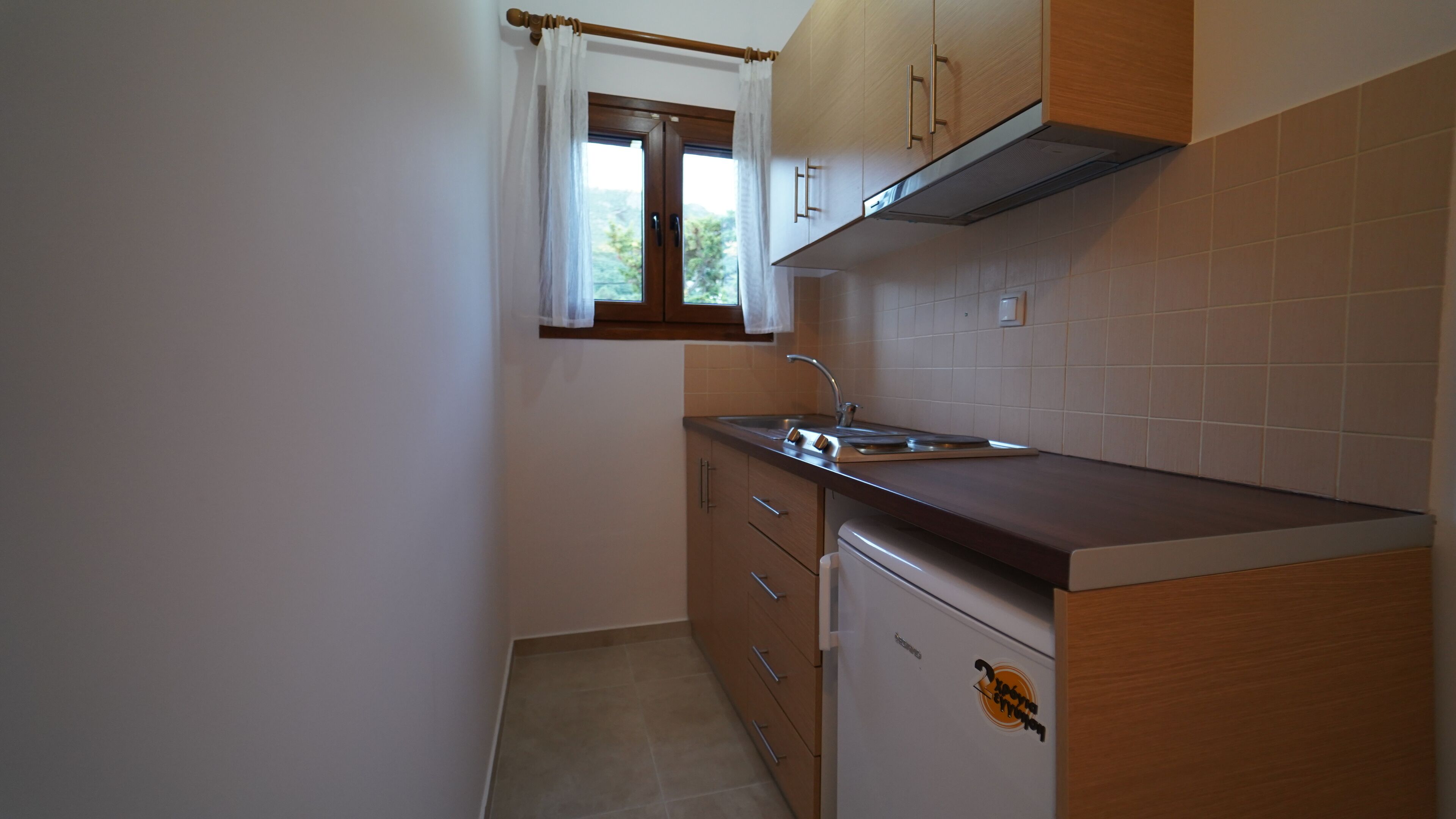 A compact kitchen with wooden cabinets, a stainless steel sink, and a window with a white curtain.