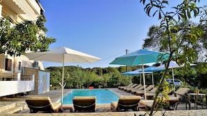 A poolside area with lounge chairs and umbrellas, a stone pool, and a building with a balcony. - Villa Apollo Parga (Parga)