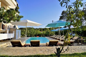 Un área junto a la piscina con sillas de descanso y sombrillas, una piscina de piedra y un edificio con balcón.