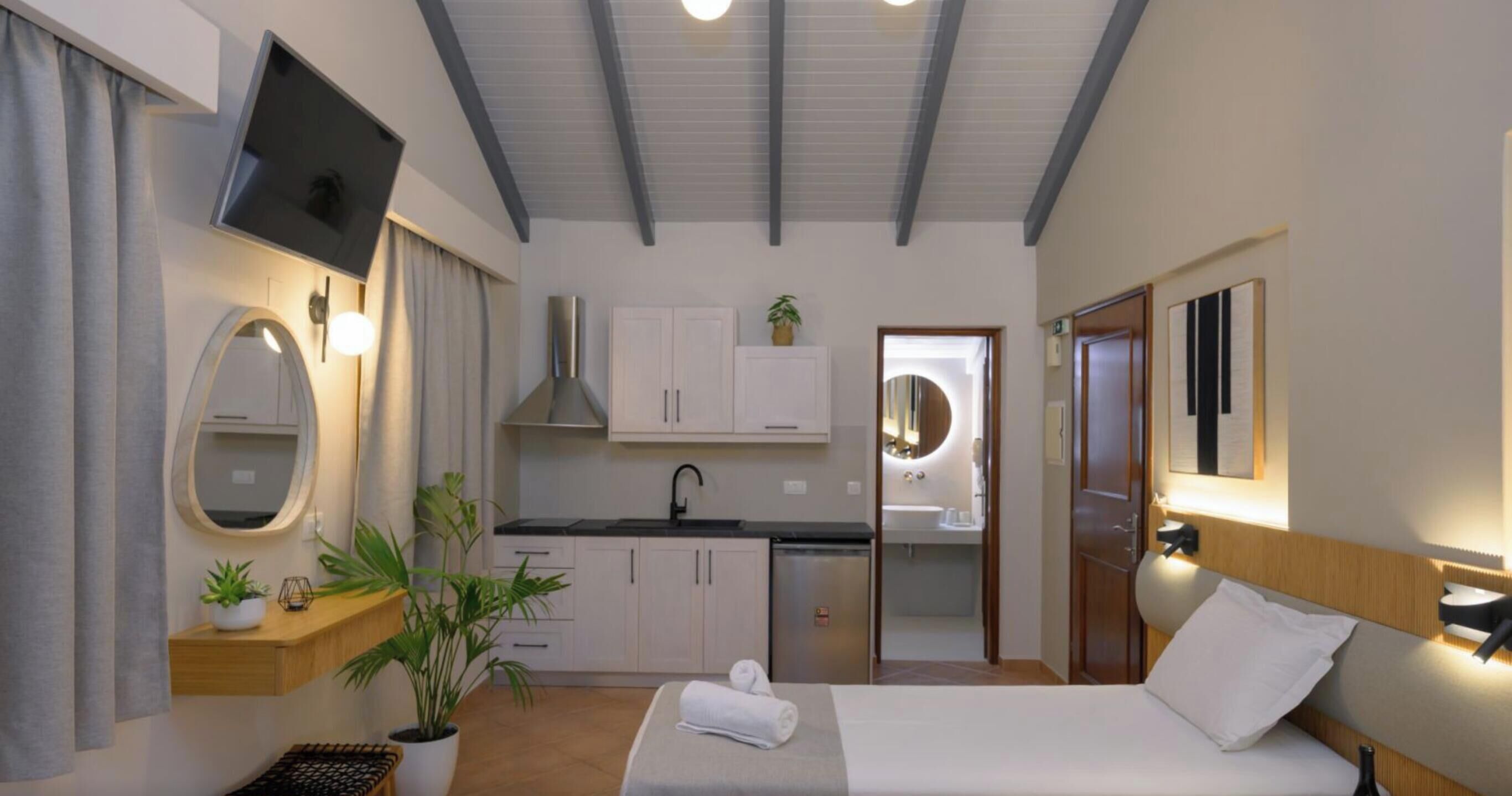 A modern hotel room with a bed, a TV, a mirror, a sink, and a small kitchen area.