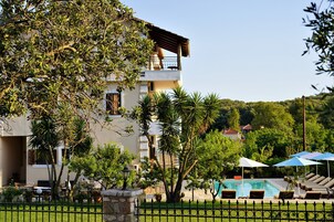 Un edificio de dos pisos con un balcón, piscina con sillas de descanso y un jardín cercado.