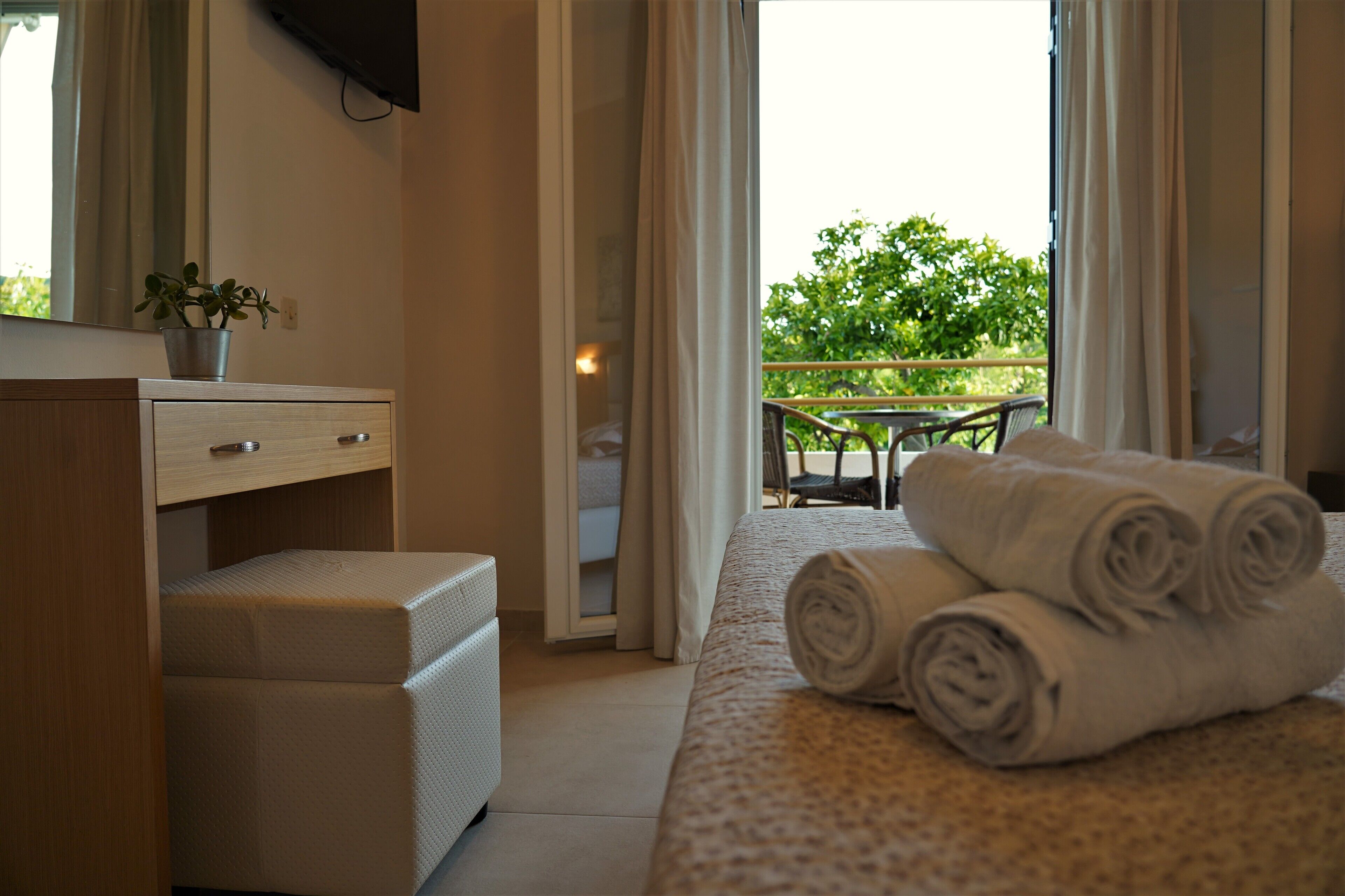 Habitación de hotel con cama, mesita de noche, escritorio con silla, planta en maceta y balcón con mesa y sillas.