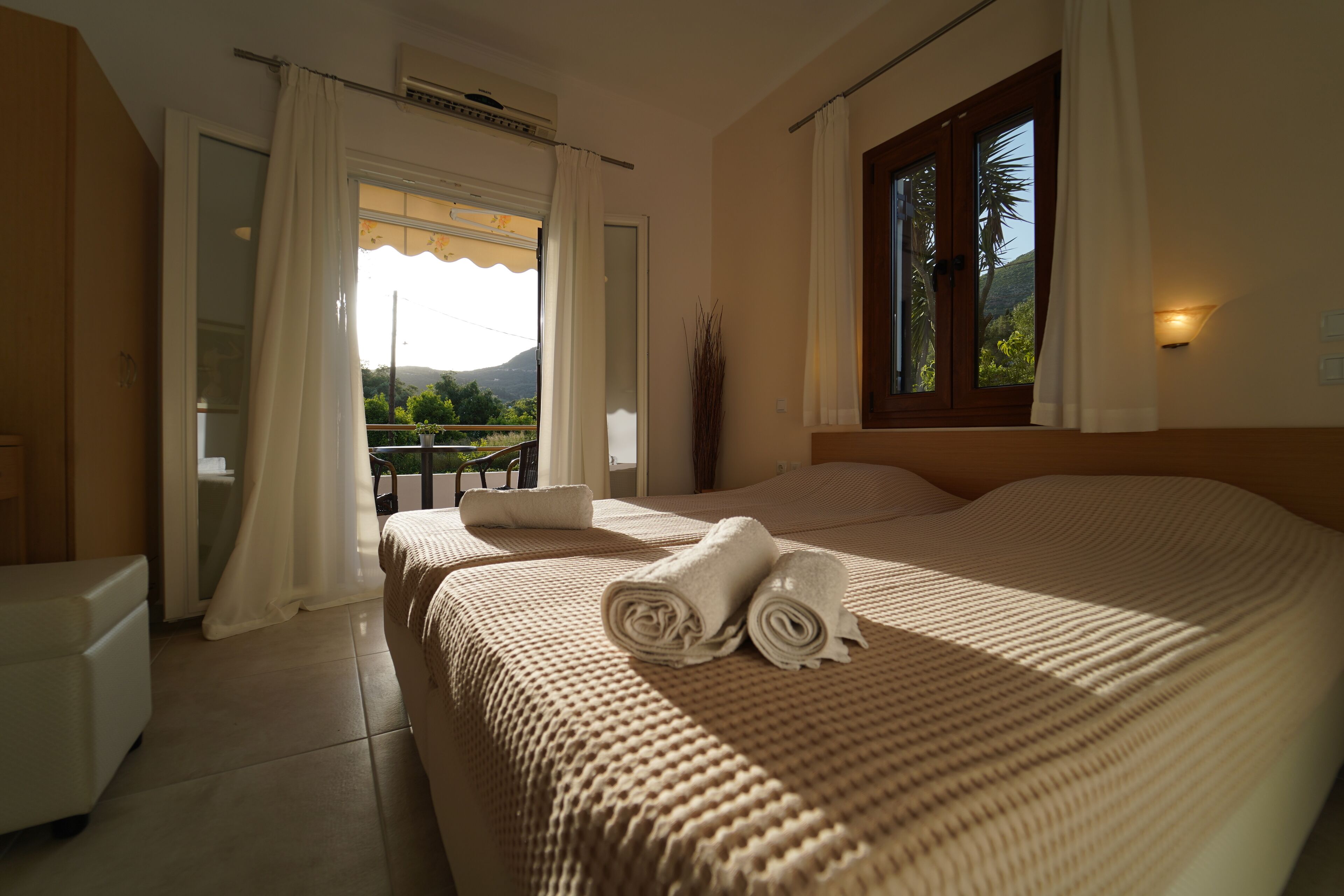 Habitación de hotel con cama, una mesita, una silla, ventana con cortinas y vista a zonas verdes y montañas.