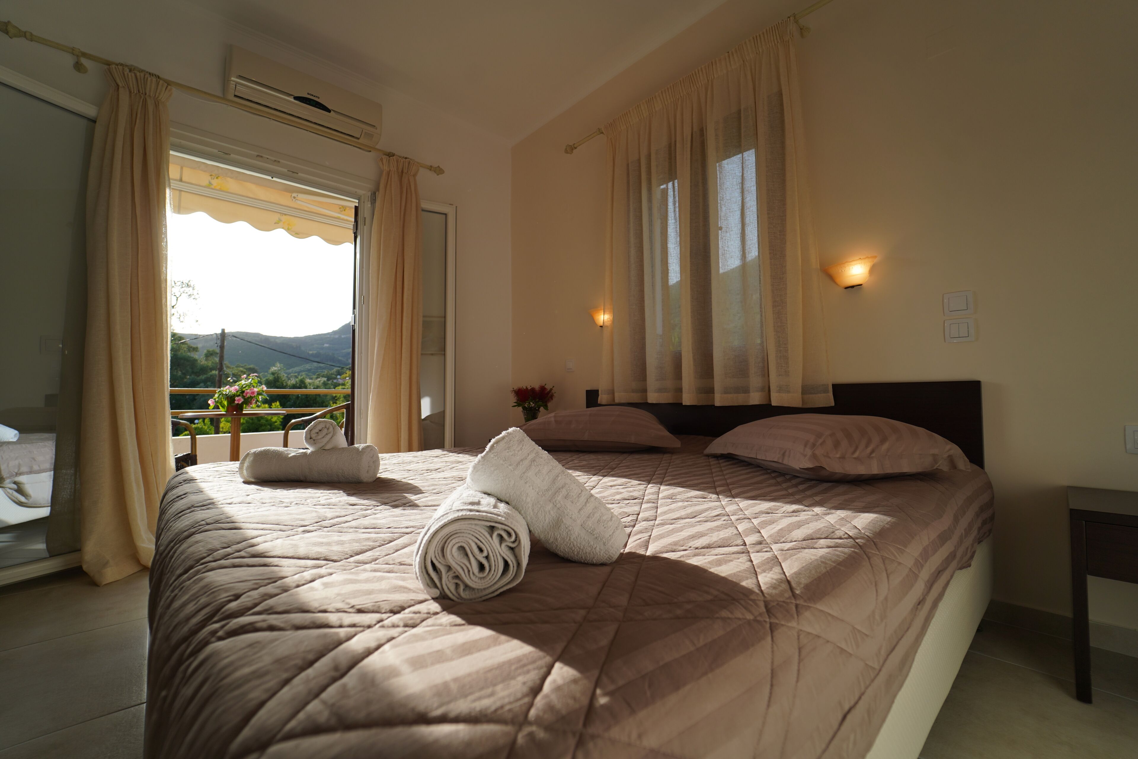 A bedroom with a large bed, a window with curtains, a bedside table, and a view of greenery outside.