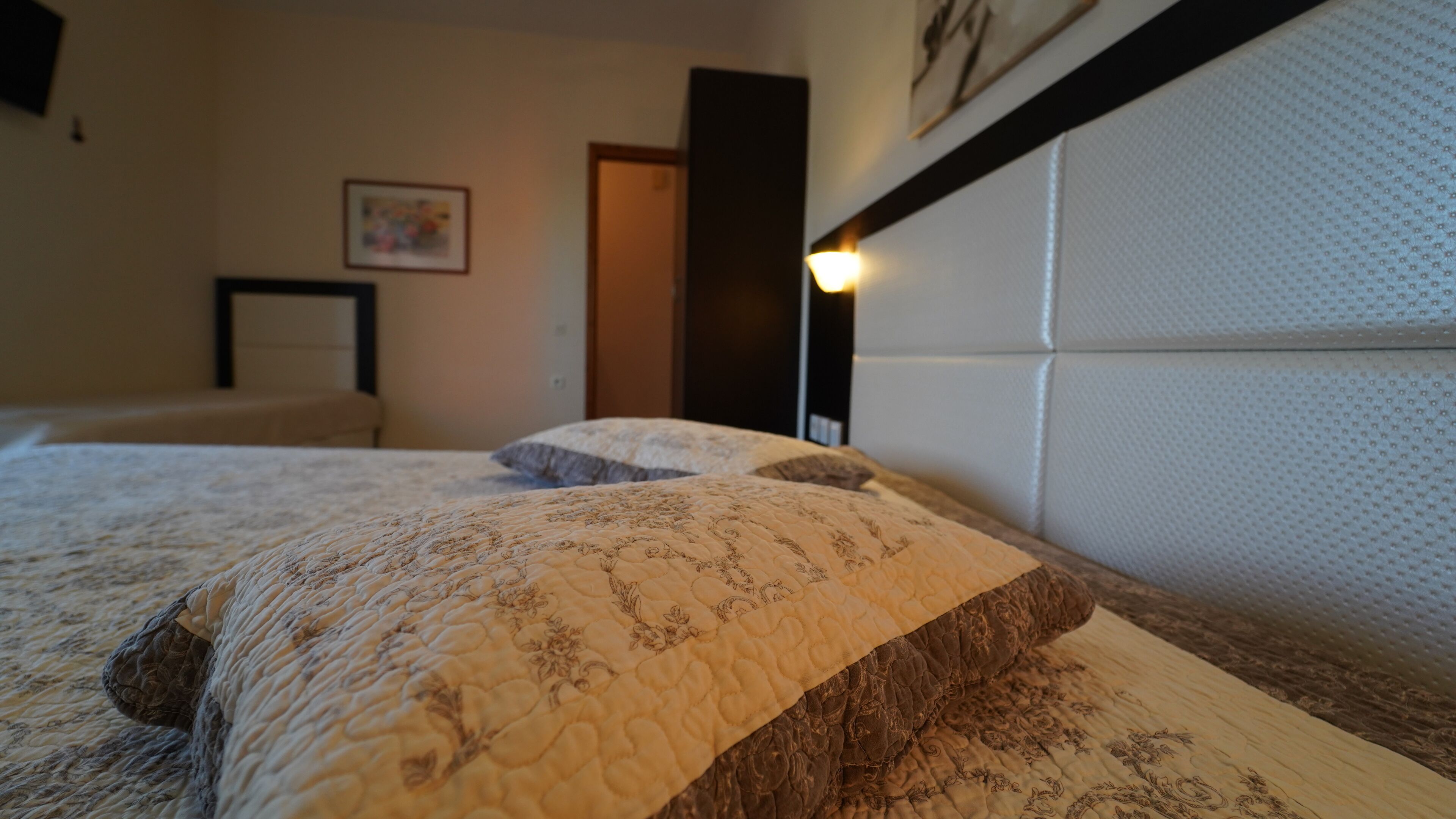 A neatly made bed with a floral-patterned bedspread and pillows, a headboard with a textured design, and a wall-mounted light fixture.