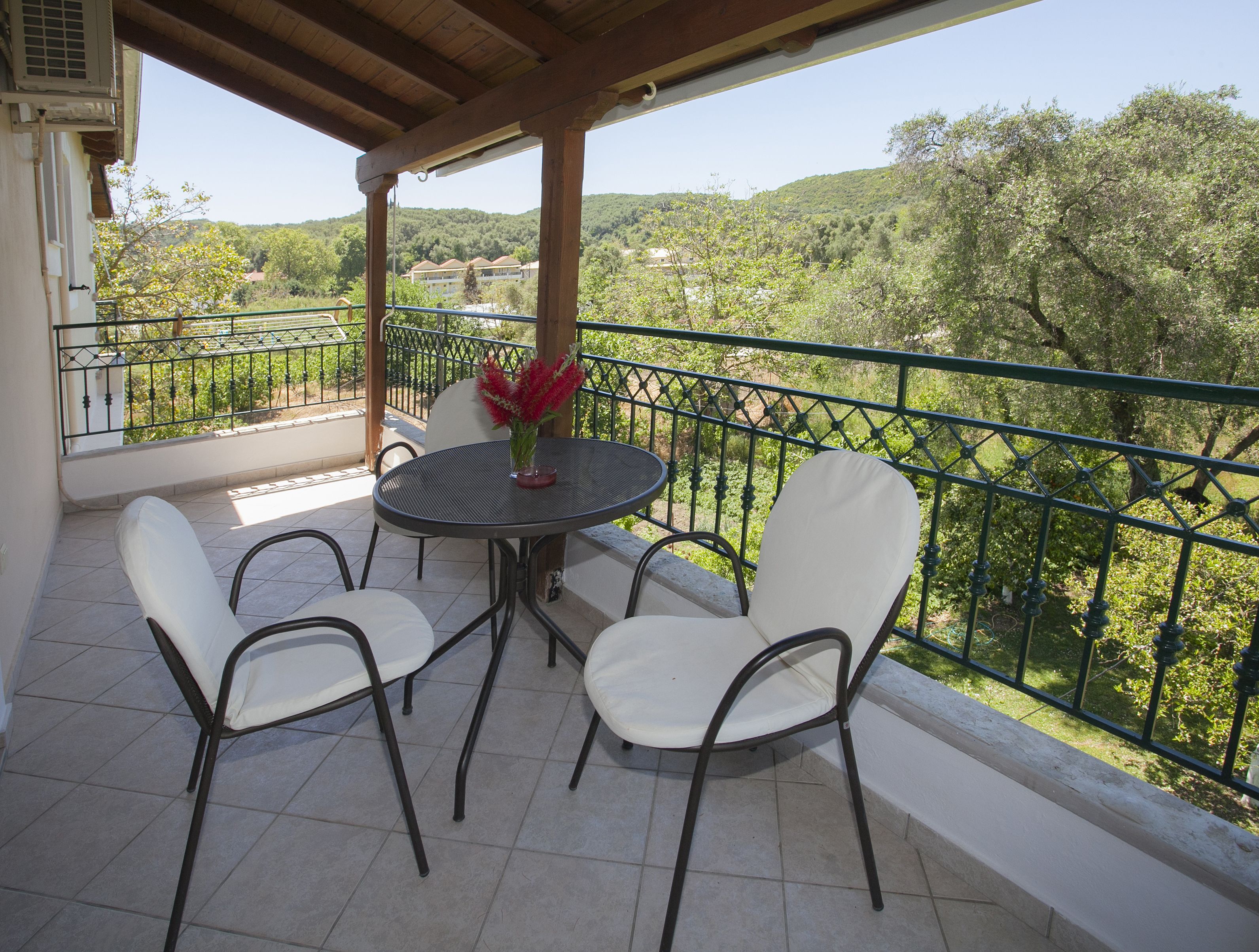 Un balcón con una mesa redonda y cuatro sillas, una planta en maceta y vistas a un paisaje verde con árboles y edificios.
