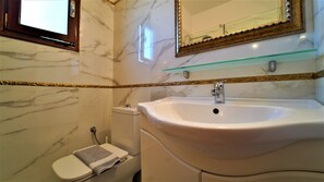 A bathroom with a marble wall, a white sink, a glass shelf above the sink, and a toilet with a towel on it.
