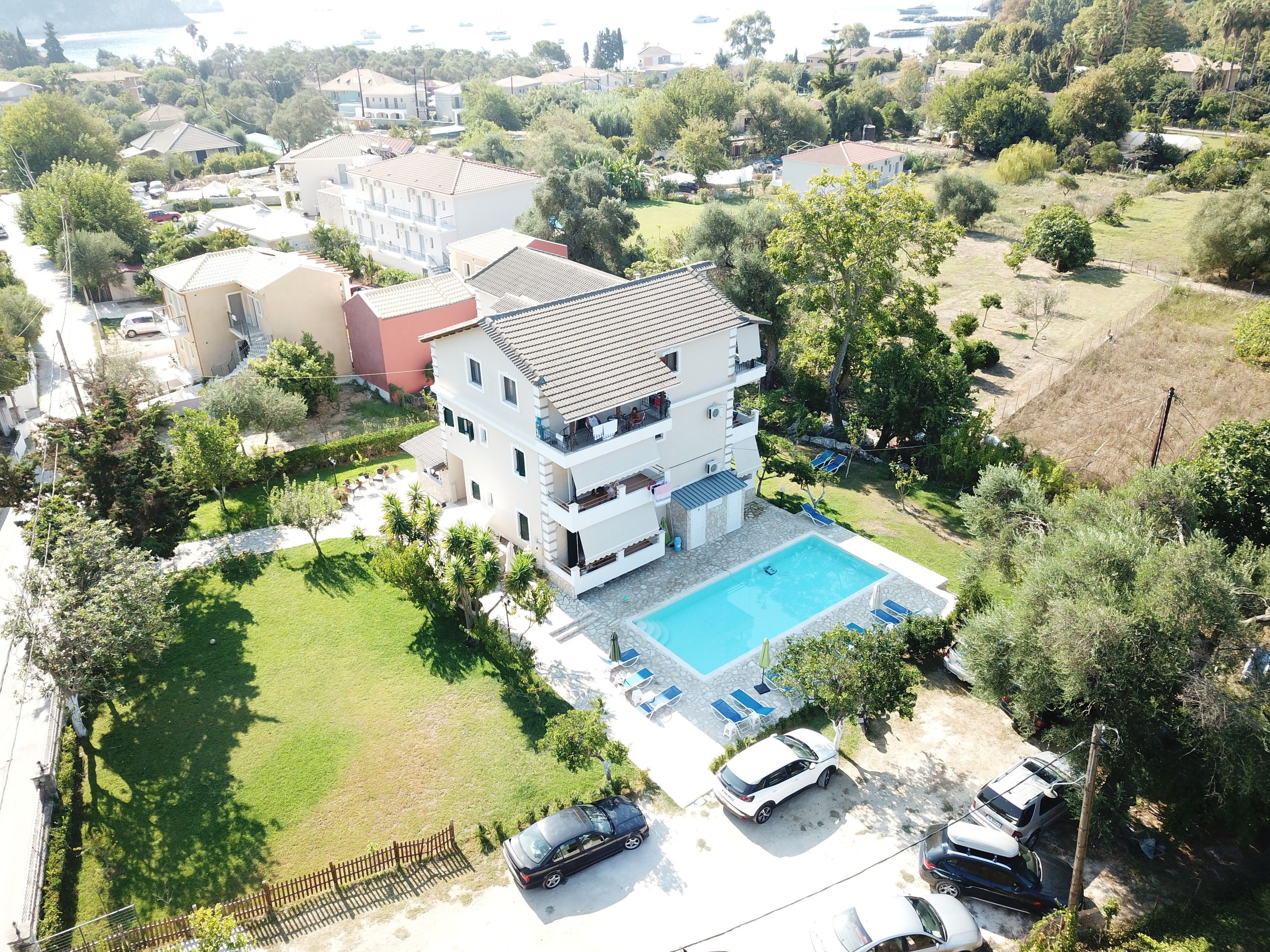 A multi-story building with a swimming pool, surrounded by greenery and parked cars.