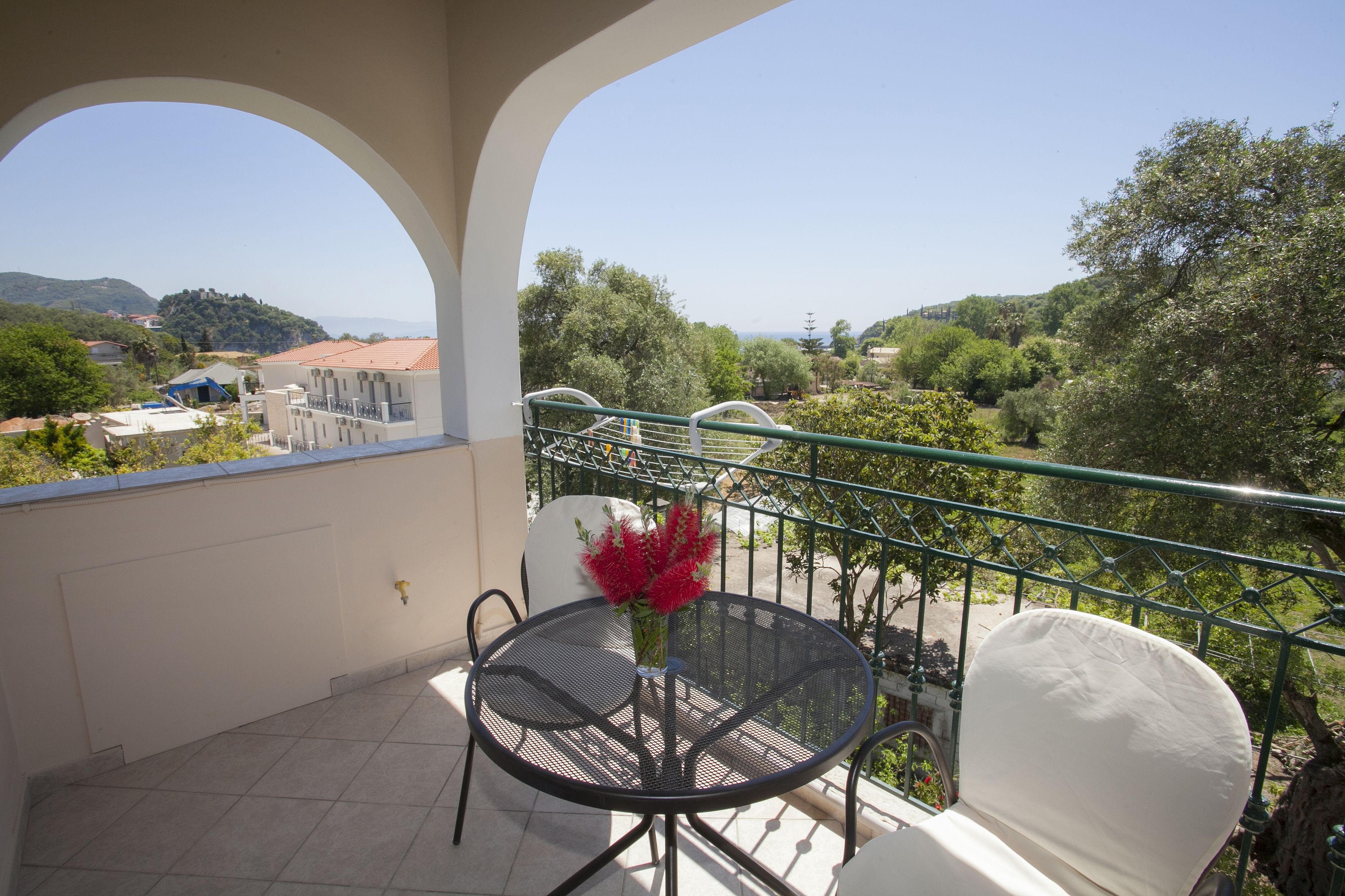 A balcony with a round table and chairs, a vase with red flowers, and a view of a green landscape with trees and buildings.