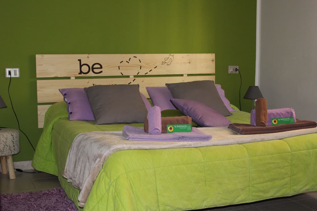 A bed with green bedding, multiple pillows, a wooden headboard with the word "be" and a drawing, and a small table with towels and a lamp.