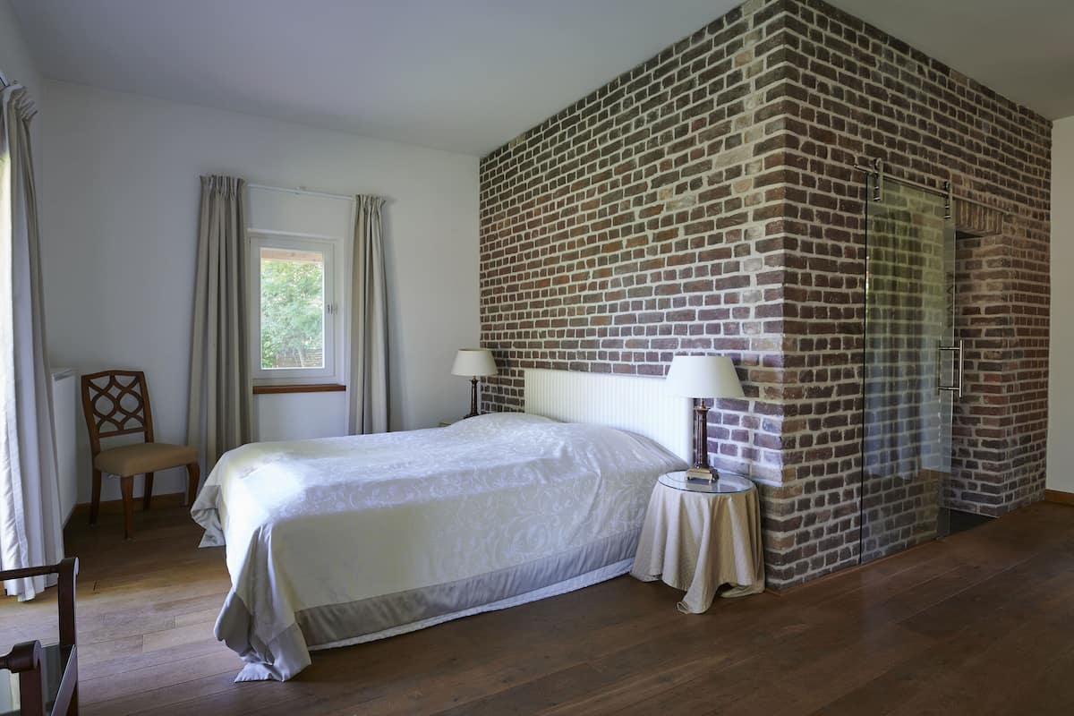 A bedroom with a brick wall, a bed, a chair, a window, and a lamp.