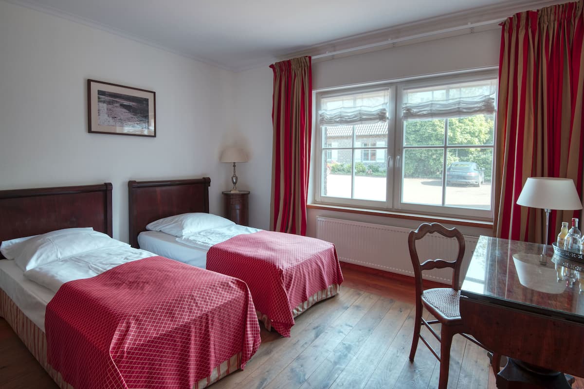 A hotel room with two beds, a wooden headboard, a table with a chair, a window with curtains, and a lamp.