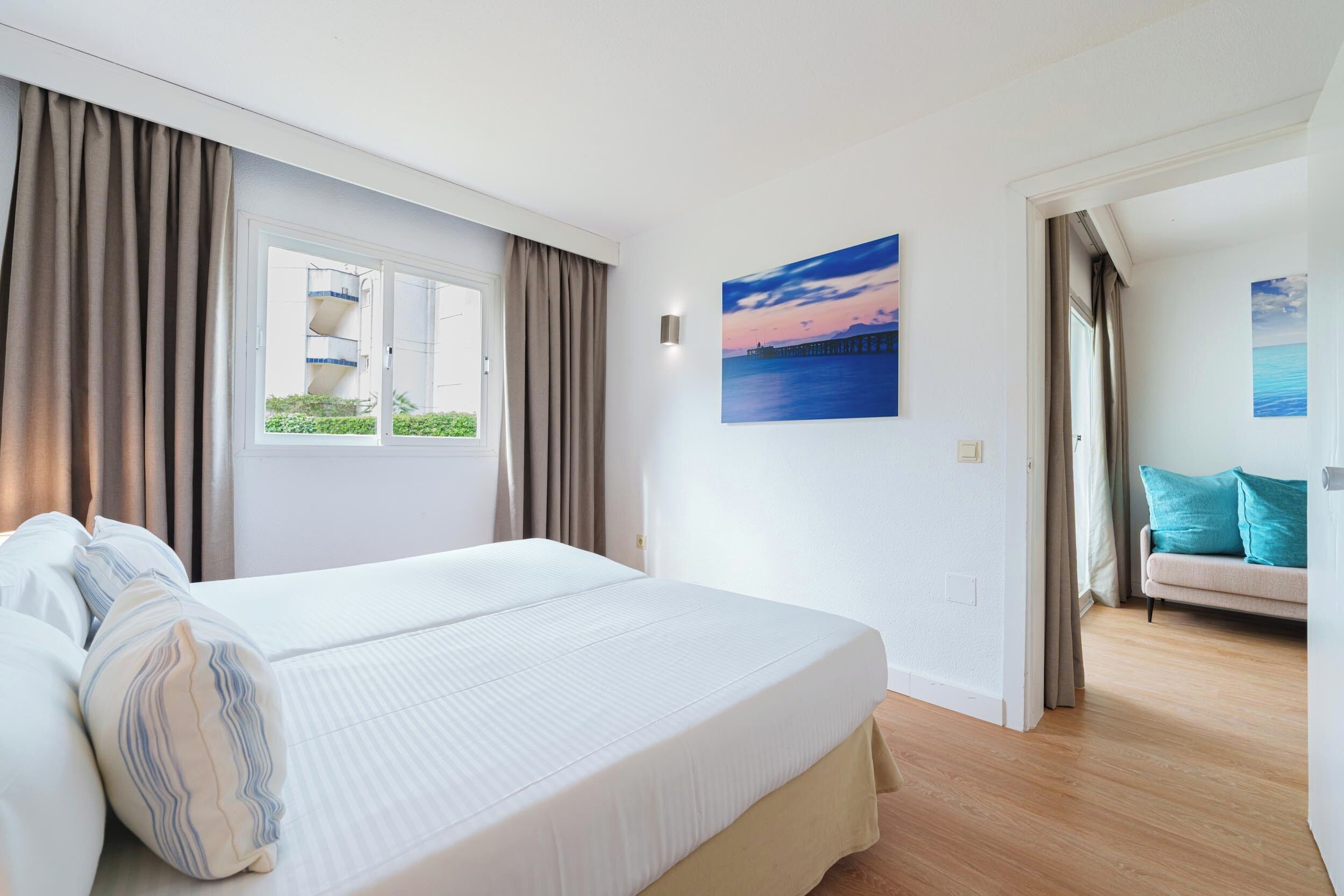 A hotel room with a large bed, a window with curtains, a painting of a sunset, and a sofa with cushions.