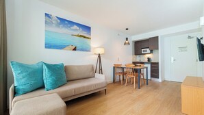 A living room with a sofa, teal cushions, a dining table with chairs, a lamp, and a kitchen area with a microwave. - Caprice Playa de Alcudia Apartments (Alcúdia)