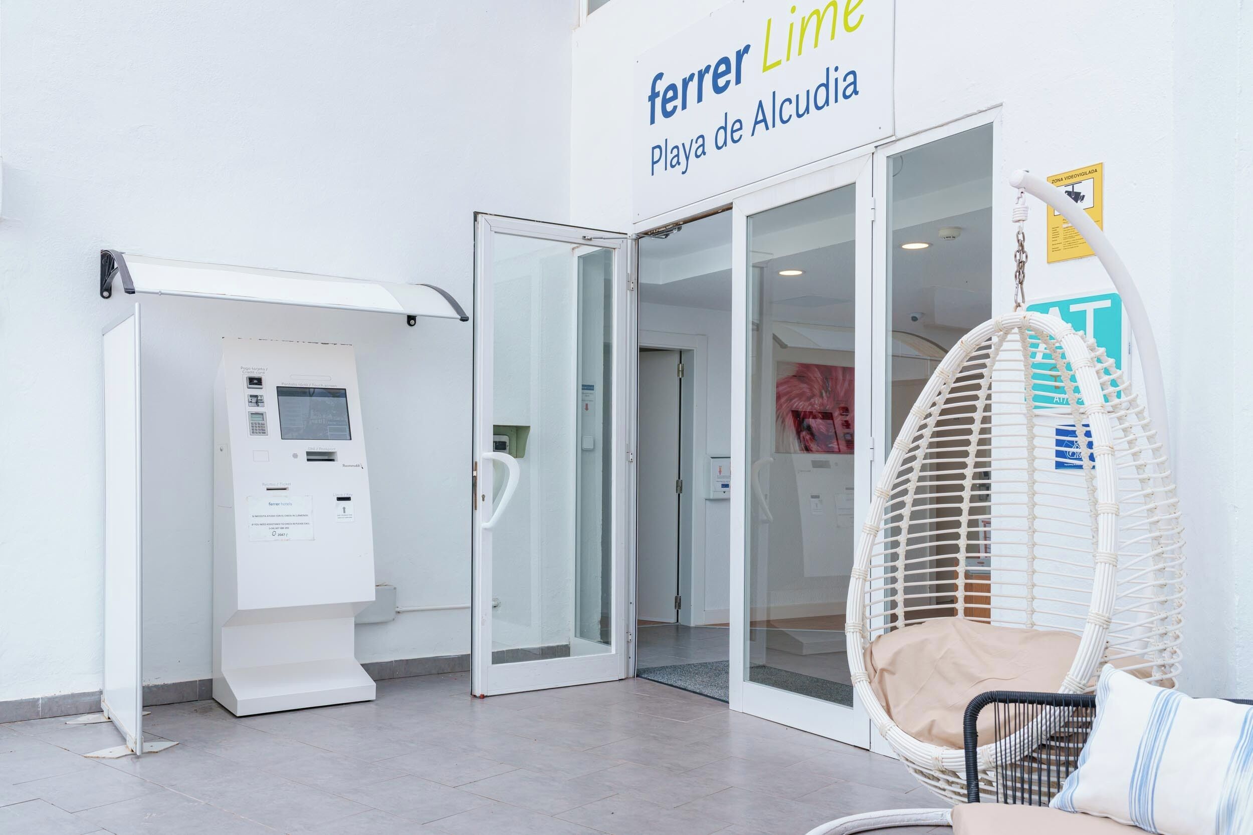 A hotel entrance with a self-service kiosk, a glass door entrance, and a wicker chair outside.