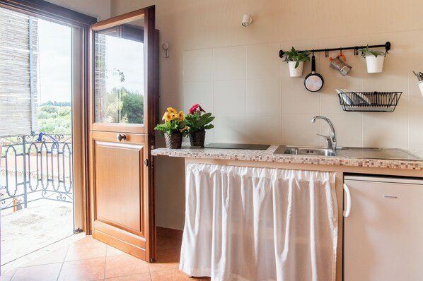 A kitchen with a sink, a dish rack, and a refrigerator. There are potted plants on the wall and a window with a view of greenery.