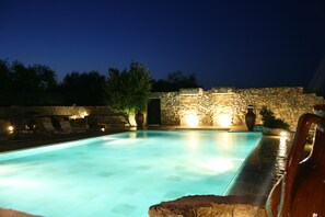 A swimming pool with stone walls and lounge chairs, surrounded by trees and a stone wall.