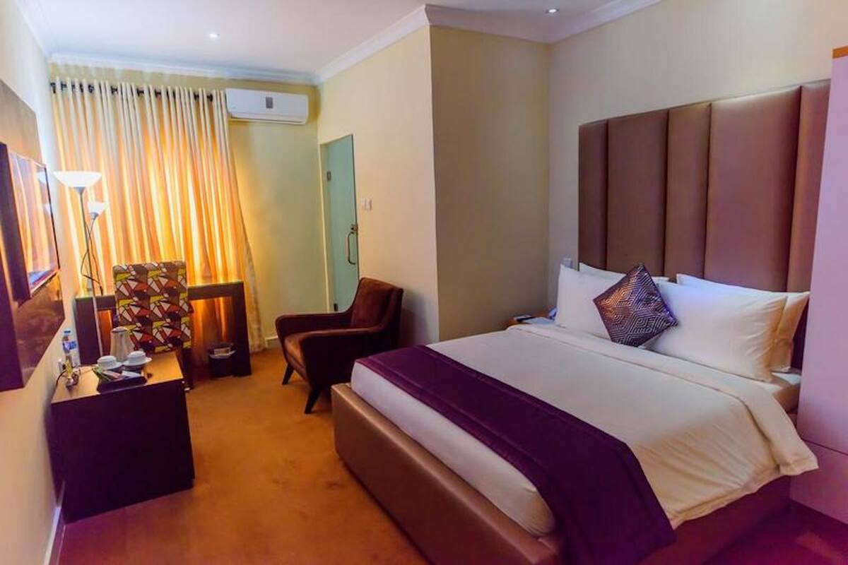 A hotel room with a large bed, a desk with a lamp, a chair, and a curtain.