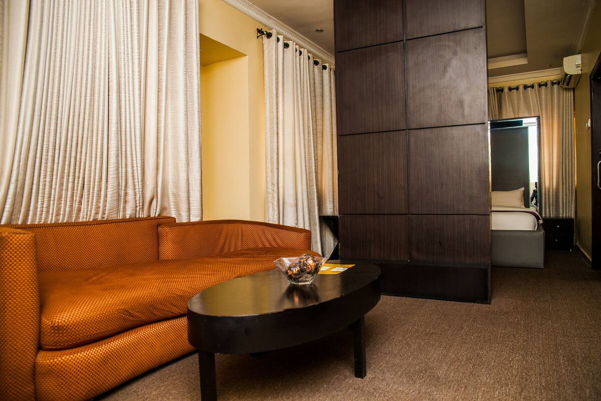 A hotel room with a sofa, a small table with a bowl, a dark wooden panel, and a bed with a white headboard.