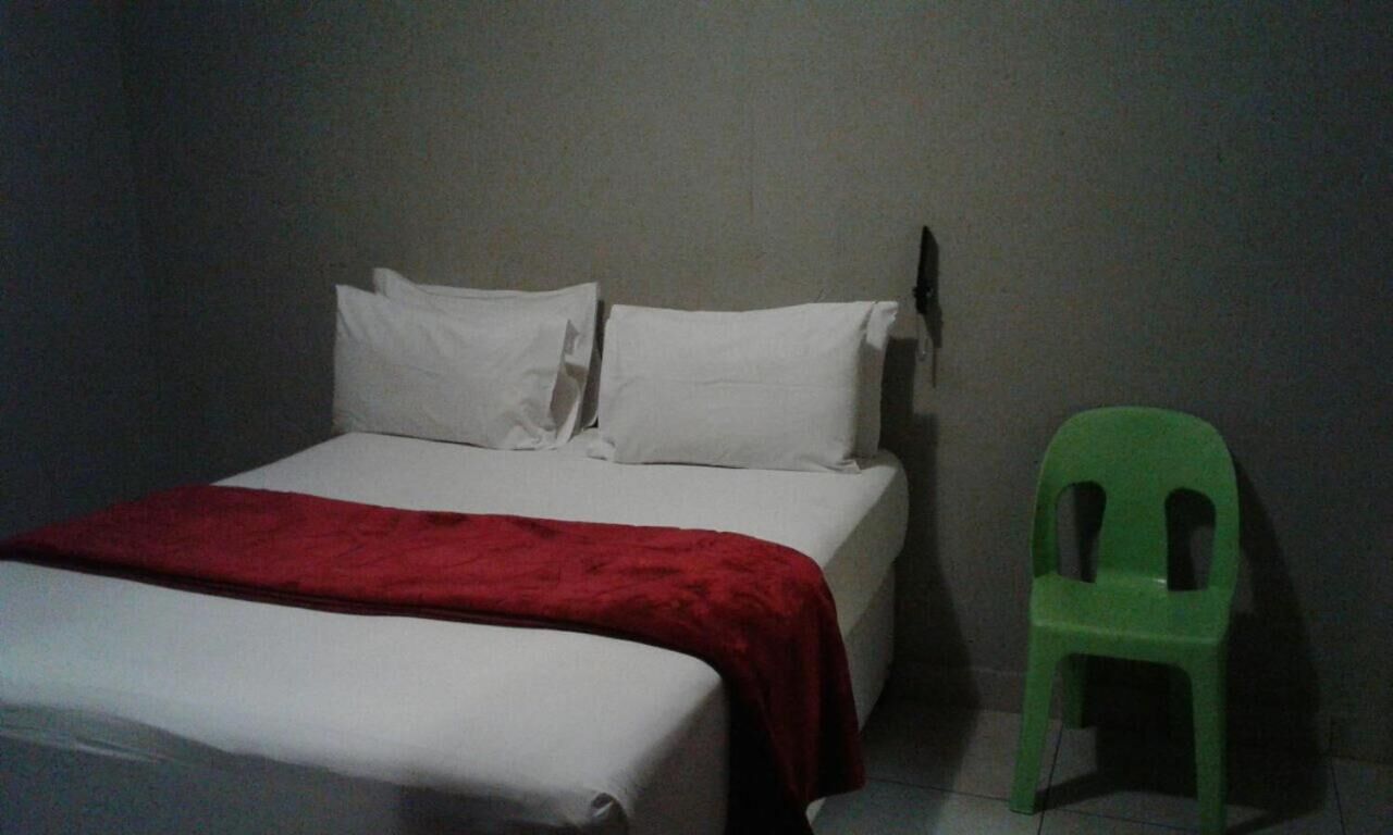 Un letto con lenzuola bianche e una coperta rossa, una sedia di plastica verde e un muro spoglio.