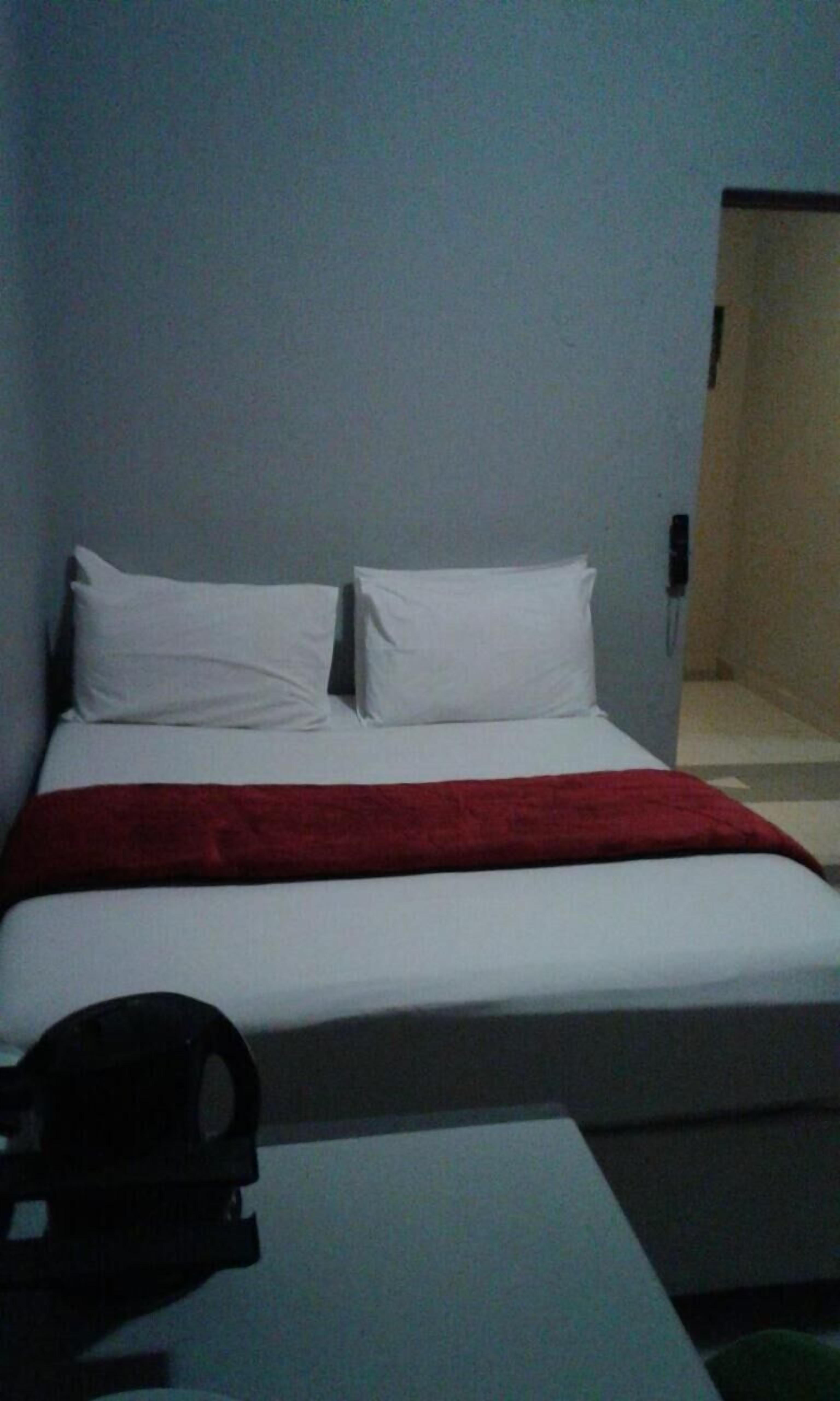 A simple hotel room with a bed, white pillows, a red blanket, and a small table with a black object on it.