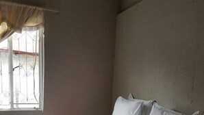 Habitación individual con una cama blanca, cabecera color beige y una ventana con cortinas.