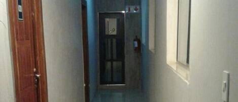 Um corredor com uma porta de madeira à esquerda, uma janela à direita e outra porta no final.