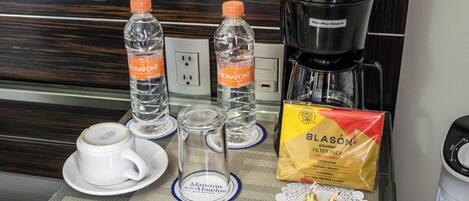 Une table de petit-déjeuner dans une chambre d’hôtel, comprenant deux bouteilles d’eau Bonafont, une machine à café, un verre, une tasse et un paquet de filtres à café Blason.