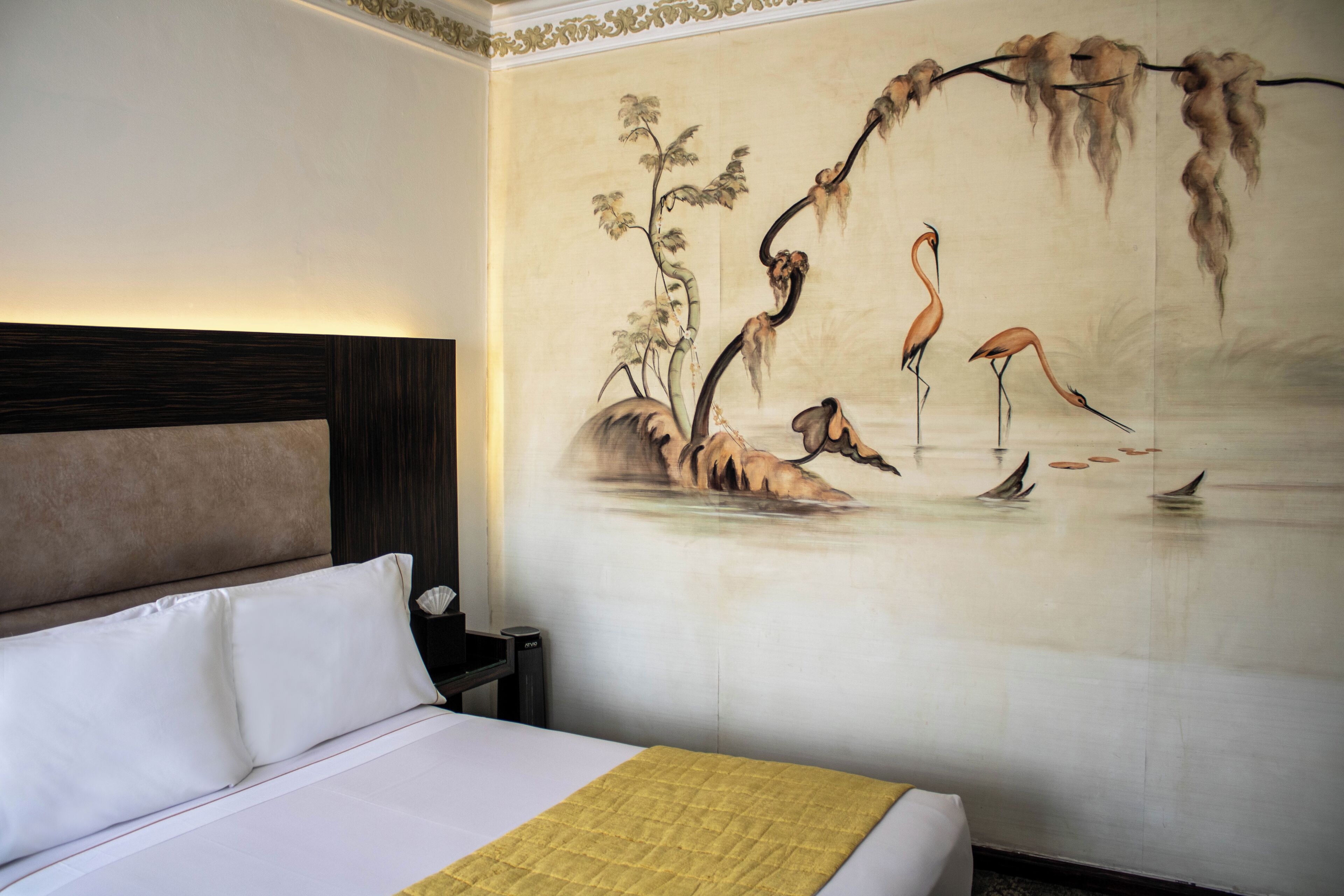 Quarto de hotel com uma cama grande, mesa de cabeceira e um mural na parede com uma árvore e flamingos.