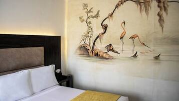 Quarto de hotel com uma cama grande, mesa de cabeceira e um mural na parede com uma árvore e flamingos.