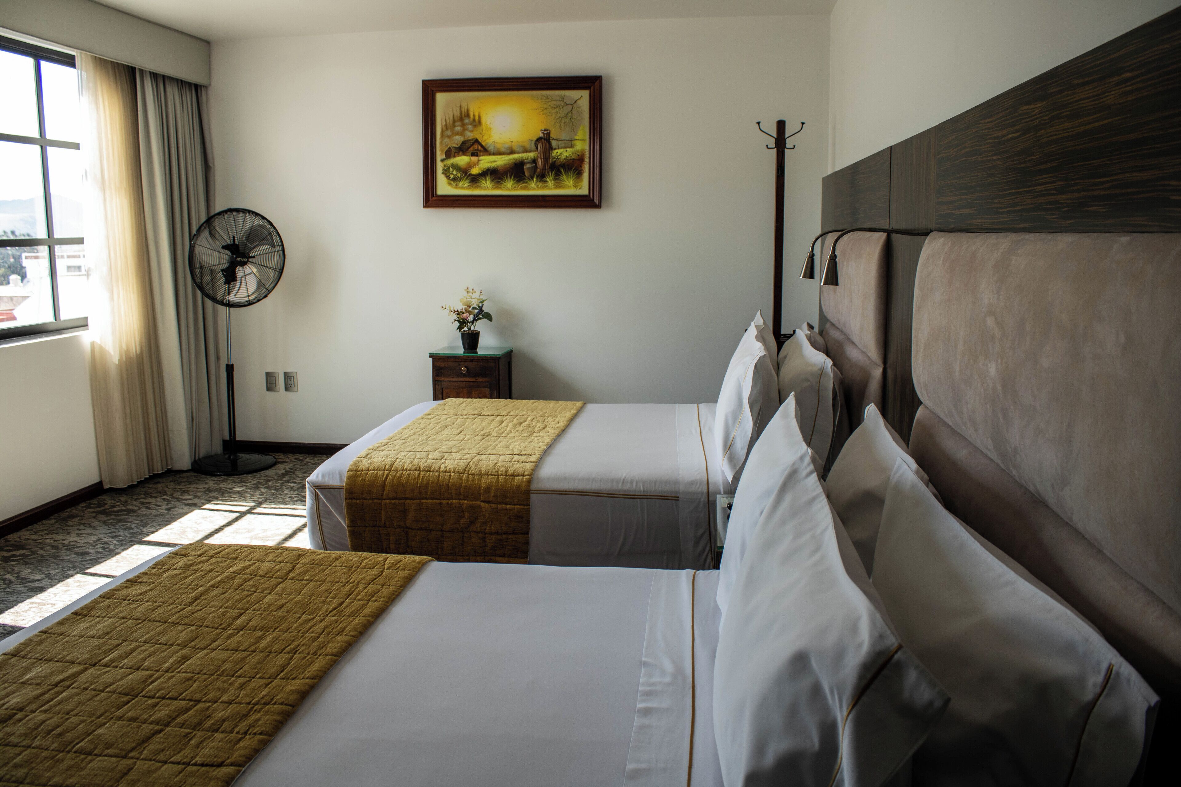 Habitación de hotel con dos camas, un ventilador, un cuadro en la pared y una ventana con cortinas.